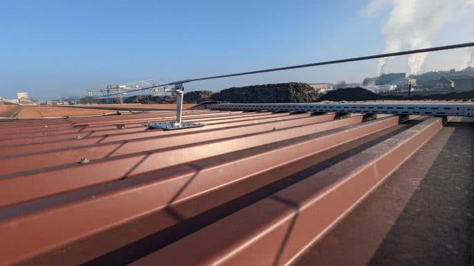 Dach mit rotem Metallbelag und Leitern, im Hintergrund ein Industriegebiet.